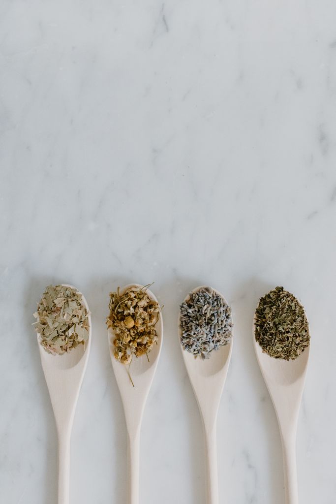 Four wooden spoons with assorted dried herbs displayed on a marble background.